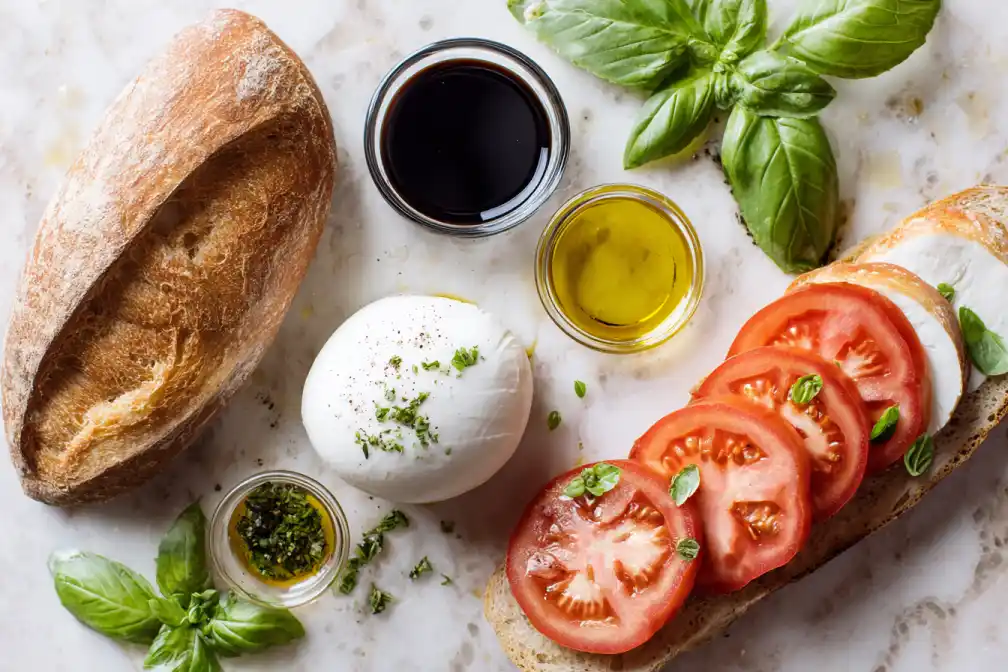 Fresh ingredients for Caprese sandwich including mozzarella, tomatoes, basil, olive oil, balsamic vinegar, and rustic bread.